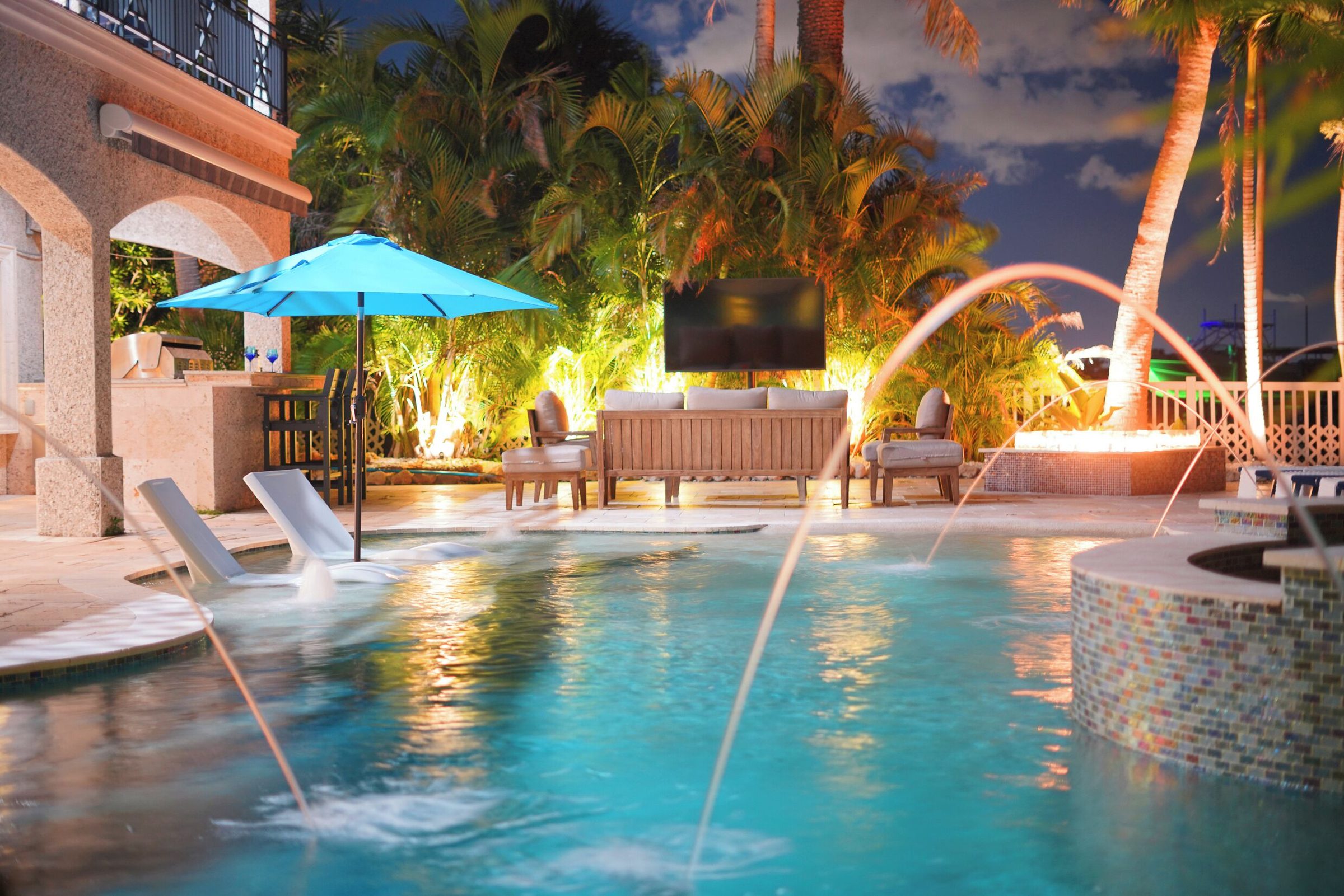 Coastal Manor pool with fountain jets, lounge chairs and an outdoor TV at dusk